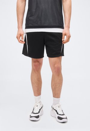 Homem vestindo calções desportivos pretos com riscas brancas, meias brancas e ténis pretos e brancos, parado contra um fundo simples.