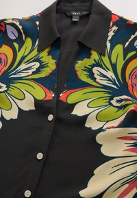 Chemise noire à boutons avec de grands motifs floraux colorés en vert, crème, bleu, rouge et orange et un col pointu noir.