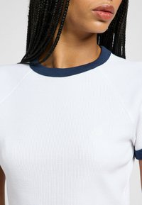 Hvid ribbet t-shirt med marineblå krave og ærmeaccent. Har et diskret brandlogo på forsiden. Teksturen virker åndbar.