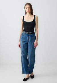 Body nero a coste con scollatura a onda, abbinato a jeans in denim blu larghi e una cintura nera; la modella indossa scarpe nere senza lacci.