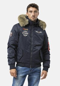 Marineblaue, wattierte Bomberjacke mit fellgefütterter Kapuze, Reißverschlusstaschen, Patches und gerippten Bündchen. Glatte Textur mit kontrastierenden Hardware-Details.