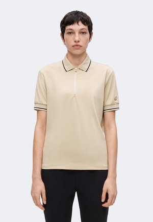 Jeune personne aux cheveux courts et foncés portant un polo beige à manches courtes avec bordure noire et un pantalon noir, debout devant un fond uni.