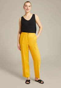Top nera senza maniche con scollo a V, abbinata a pantaloni in lino gialli a vita alta con un bottone frontale e due tasche laterali. Sandali neri.