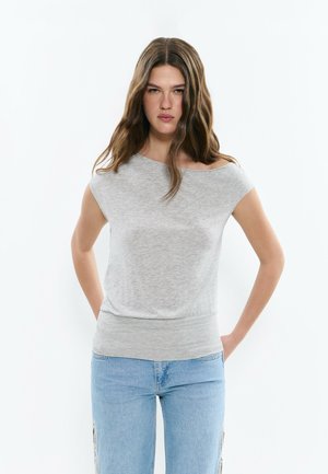 Joven con cabello largo y castaño, vestida con una blusa gris claro sin hombros y vaqueros azul claro, de pie con las manos detrás de la espalda.