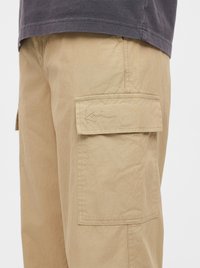 Gros plan sur un pantalon cargo beige avec une poche à rabat sur le côté présentant un détail de signature cousu et une chemise gris foncé par-dessus.