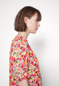 Blusa floral com um estampado colorido de flores rosas, amarelas e verdes sobre um fundo claro, com mangas curtas bufantes e um corte descontraído.