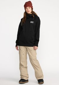 Sweat-shirt à capuche noir avec une poche avant et un logo blanc. Pantalons cargo beiges avec des zips et des poches, associés à des bottes noires et un bonnet rouge à motifs.