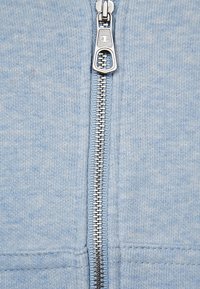 Ljusblå zip-hoodie med silverdragkedja, ribbad textur och kontrasterande sömnadsdetaljer. Dragkedjan har en rund dragflik.