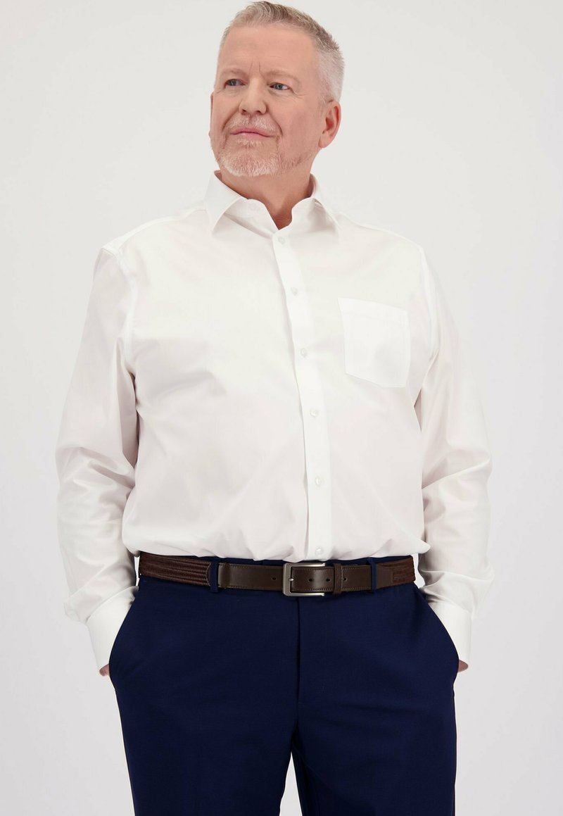 Middelgrote man met kort grijs haar, gekleed in een wit overhemd met knopen en marineblauwe broek, met zijn handen in zijn zakken, die iets opzij kijkt.