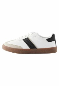 Białe sneakersy z czarnymi paskami, gumową podeszwą i perforowanymi akcentami. Posiadają wiązany design oraz kontrastującą beżową osłonę palców.