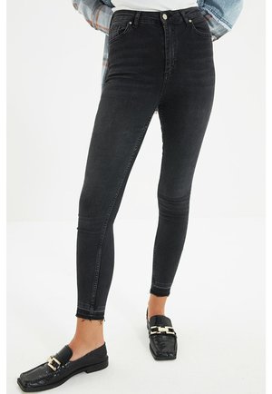 Person trägt schwarze, hoch taillierte Skinny Jeans mit rohen Säumen und schwarze Loafers mit goldenen Schnallendetails, steht vor weißem Hintergrund.