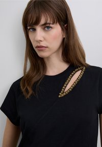 Black t-shirt with short sleeves, featuring a decorative gold embellished opening on the shoulder. Smooth texture and casual design.