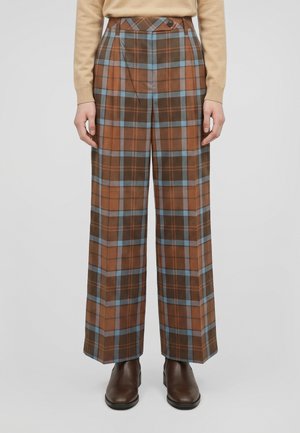 Modèle portant un pantalon marron à carreaux, à jambes larges et à rayures bleues et orange, un pull beige et des bottines marron.