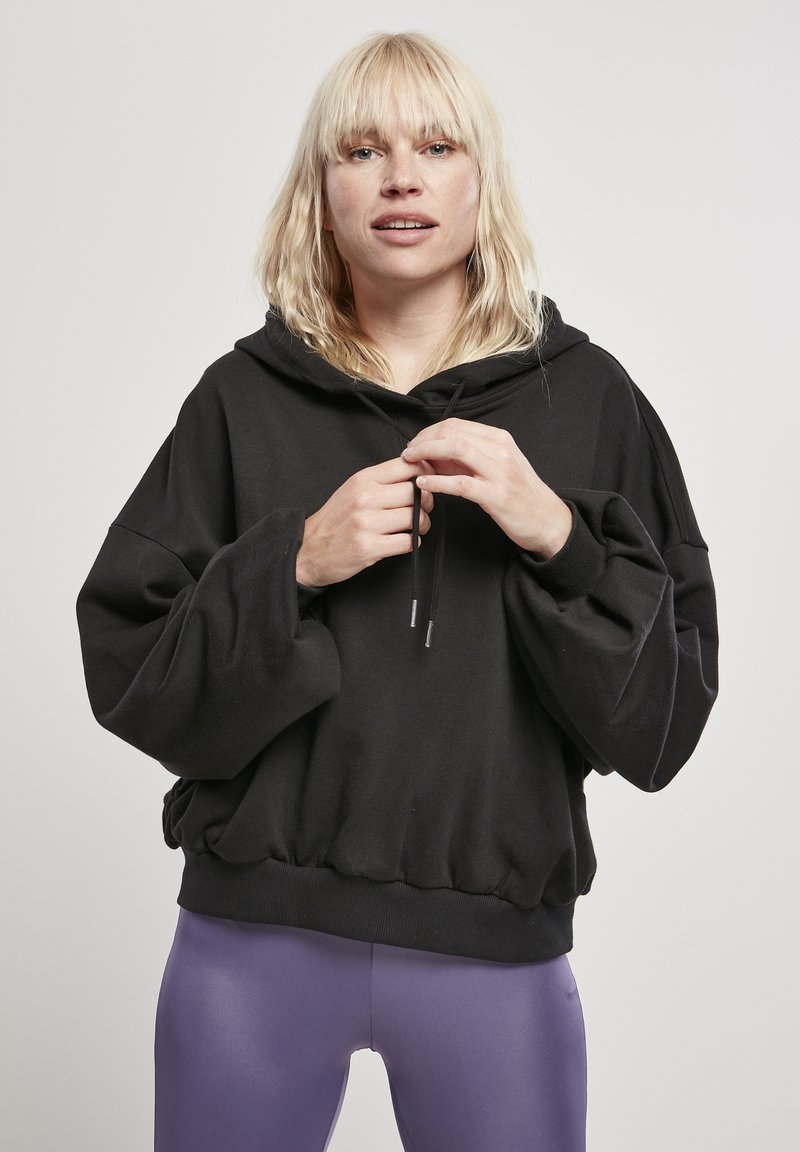 Felpa nera oversize con cappuccio regolabile, maniche larghe e orlo a coste. Indossata sopra leggings viola, caratterizzati da una texture liscia e elastica.