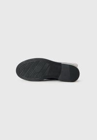 Semelle de chaussure en caoutchouc noir avec un motif texturé, comportant des rainures pour l'adhérence et une légère élévation du talon. Dessous propre, design minimaliste.