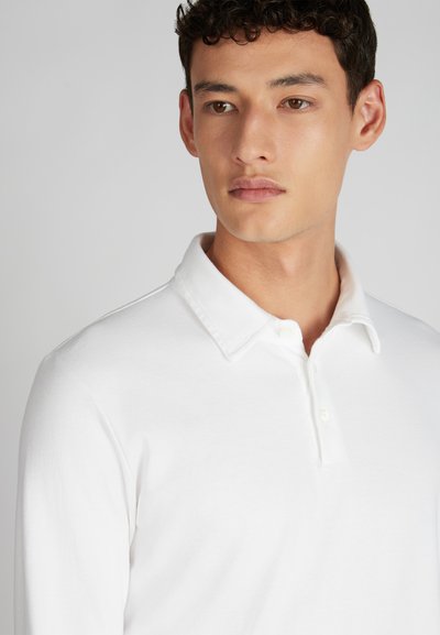 Giovane uomo con i capelli corti e scuri, che indossa una polo a maniche lunghe bianca, che guarda leggermente di lato su uno sfondo neutro.