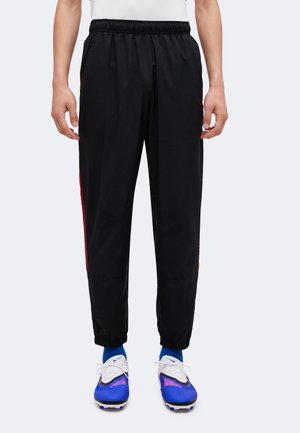 Homme portant un pantalon de jogging noir à taille élastique avec des bandes rouges sur les côtés, des chaussettes bleues et des crampons bleus et blancs, debout devant un fond uni.
