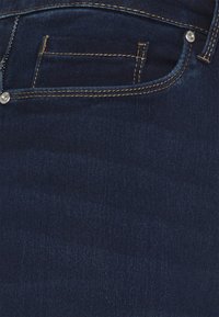 Gros plan sur un tissu en denim bleu foncé montrant une poche avant avec une petite poche à monnaie et des coutures visibles avec des rivets métalliques.