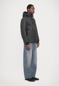 Schwarze, gesteppte Pufferjacke mit Kapuze, die eine strukturierte Oberfläche und elastische Bündchen aufweist, kombiniert mit hellblauen, weiten Jeans und schwarzen Stiefeln.