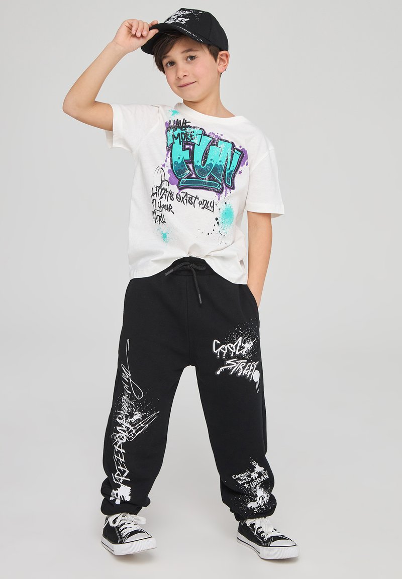 T-shirt grafica bianca con scritte in stile spray blu e viola, abbinata a pantaloni neri con accenti di graffiti bianchi. Cappellino nero e scarpe da ginnastica.