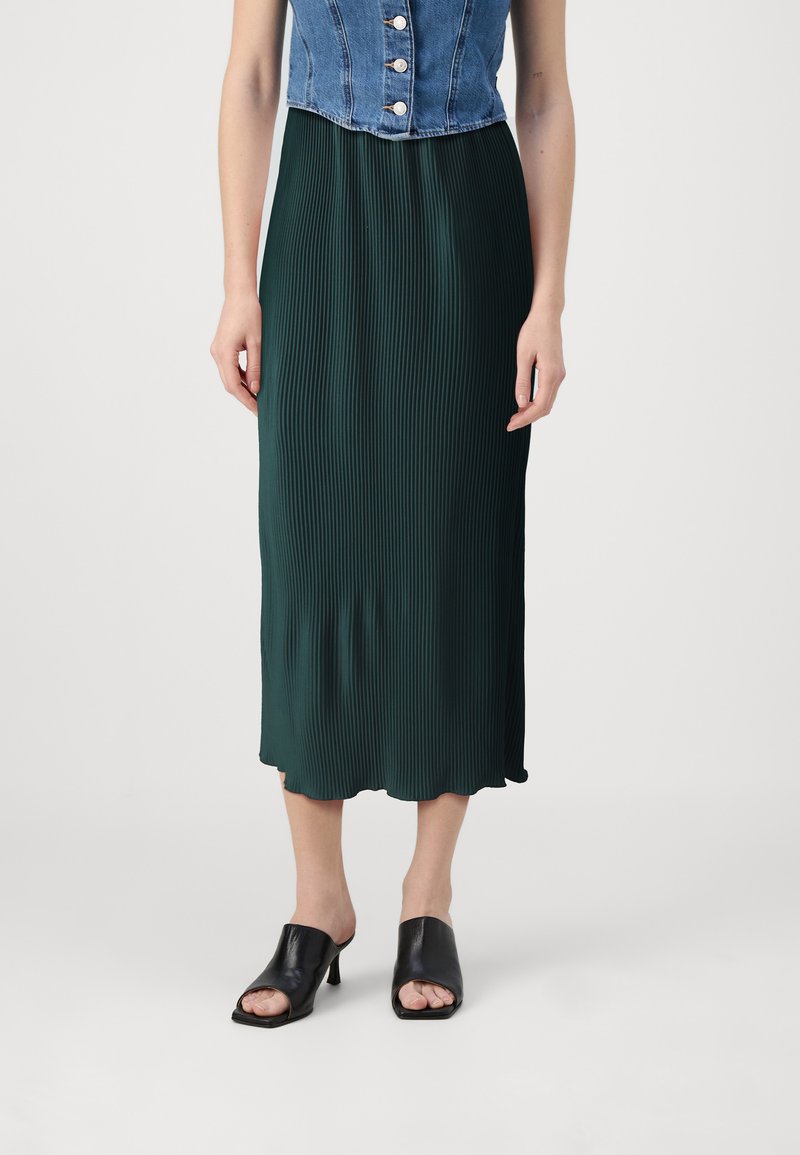 Fustă maxi plisată verde cu margine ondulată, asortată cu un top din denim scurt și mule negre cu vârful deschis. Textură netedă și design fluid.