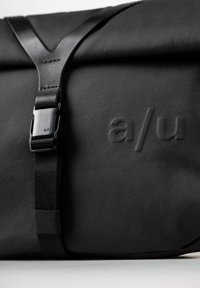 Borsa a tracolla nera con finitura opaca, dotata di una patta pieghevole e una distintiva fibbia nera. Il logo "a/u" è impresso sulla superficie.