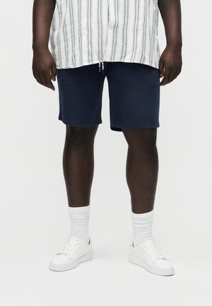 Vyras, dėvintis tamsiai mėlynas šortus, baltą dryžuotą marškinius, baltas kojines ir baltus sportbačius, stovintis priešais paprastą šviesų foną.