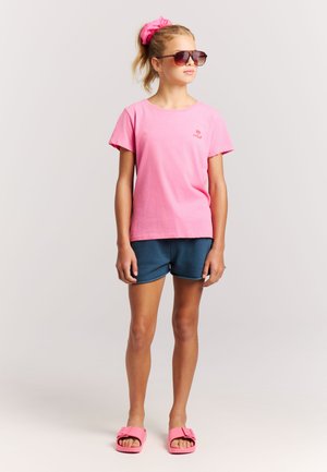 Roze katoenen t-shirt met korte mouwen, blauwe korte broek en roze slippers. De look wordt compleet gemaakt met een bijpassende roze haargom en een zonnebril.
