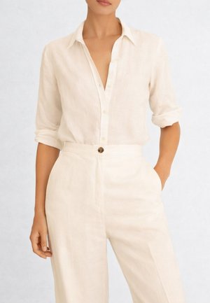 Donna che indossa una camicia color crema in lino con le maniche arrotolate, infilata in pantaloni color crema a vita alta con bottoni, con una mano in tasca.