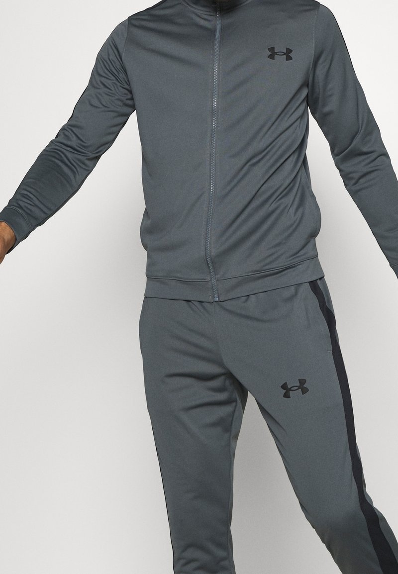 Homme portant une veste de sport zippée gris foncé et un pantalon assorti avec des logos Under Armour noirs et des bandes latérales.