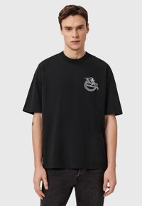 Μαύρο βαμβακερό t-shirt με κοντά μανίκια, στρογγυλή λαιμόκοψη και ένα μπροστινό γραφικό με έναν δράκο περικυκλωμένο από κείμενο. Ελαφρώς χαλαρή εφαρμογή.