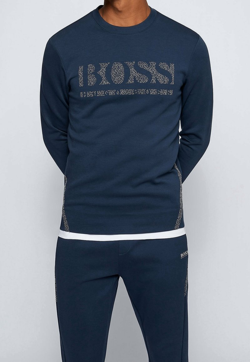 Man wearing navy blue long-sleeve shirt and pants with dotted "BOSS" logo on the chest and left thigh, standing with hands behind back.
