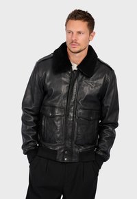 Černá kožená bombarďáka s beránkovým límcem, předními kapsami se zmáčknutím a hřebíkovými manžetami. Hladká textura, klasický design.
