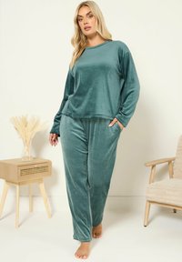 Femme en tenue de détente en velours teal avec des manches longues et un pantalon, debout pieds nus près d'une table d'appoint en bois et d'un fauteuil beige.