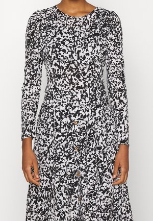 Langærmet sort-hvid abstrakt print kjole med knapper ned foran og figursyet talje, båret af en person med brun hud.