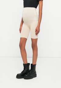 Beiga graviditetsshorts i stretchigt material, med hög midja och slät yta, kombinerade med svarta snörkängor.