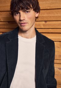 Dark corduroy blazer over a light beige t-shirt, featuring a structured collar and textured fabric. Background of wooden slats.