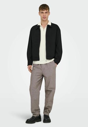 Jaunas vyras dėvi juodą striukę, kreminį pusiau užtrauktuko megztinį, pilkas kelnes ir juodus batus, stovėdamas su viena ranka kišenėje.