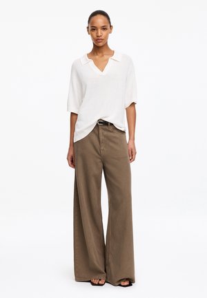 Femme portant un t-shirt blanc à manches courtes et col en V, rentré dans un pantalon marron taille haute à jambes larges, et des sandales noires à bout ouvert.