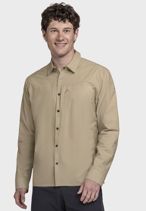 Junger Mann mit lockigem Haar, der ein beiges Hemd mit Knöpfen und dunkle Hose trägt, lächelnd und vor einem schlichten hellen Hintergrund steht.