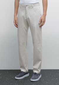 Light beige cotton trousers with a straight cut, zip fly, single button closure, and two front pockets, paired with gray suede shoes.