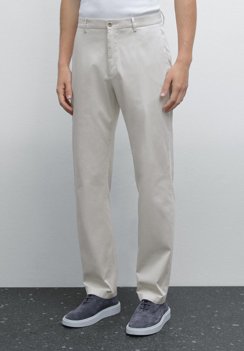 Light beige cotton trousers with a straight cut, zip fly, single button closure, and two front pockets, paired with gray suede shoes.