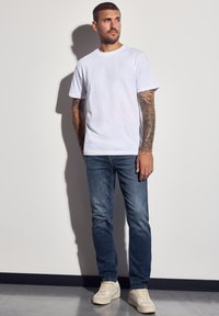 Vit bomulls t-shirt med korta ärmar, tillsammans med blå denimjeans. Modellen bär ljusa sneakers; armarna är prydda med intrikata tatueringar.