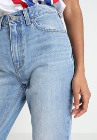 Ljusblå högmidjade jeans med en klassisk denimstruktur, utrustade med framfickor och stängning med metallknapp, delvis synlig hand.