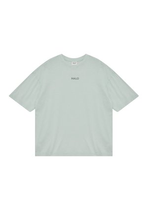 Mężczyzna stoi, ubrany w oversize'ową koszulkę w kolorze szałwii z napisem "HALO", szare szorty, białe skarpetki i szare trampki, zwrócony przodem.