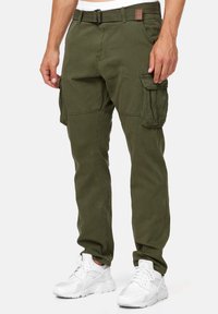 Oliivinvihreät cargo-housut, valmistettu puuvillasta, joissa on useita taskuja, suora leikkaus ja säädettävä vyötärö. Yhdistetty valkoisiin lenkkareihin.