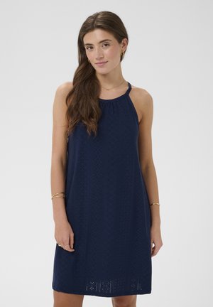 Femme aux longs cheveux bruns portant une robe dos nu bleu marine sans manches et des bijoux en or, debout devant un fond clair uni.