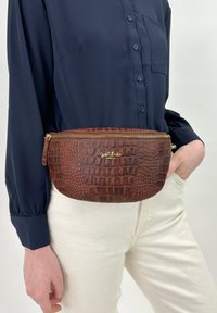 Marsupio in pelle marrone con effetto coccodrillo, dotato di zip dorata e con un logo sul davanti. Indossato in vita, abbinato a una blusa blu navy e pantaloni bianchi.