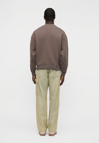 Καφέ βαμβακερό φούτερ με ριμπ μανσέτες, συνδυασμένο με ανοιχτό πράσινο τζιν παντελόνι φαρδιάς γραμμής με πίσω τσέπες και λεπτομέρεια πλαϊνού φερμουάρ.