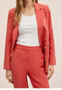 Coral blazer with two buttons, notched lapels, and a textured finish, paired with matching trousers. White tank top underneath.